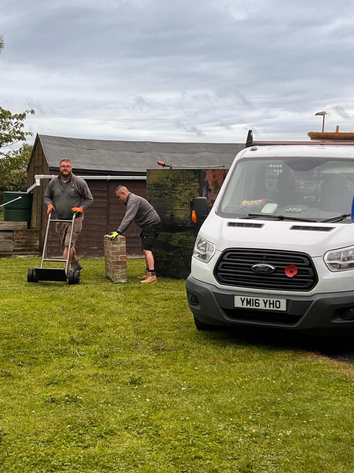 Two engineers removing an old black steel oil tank from a garden on a sack truck, alongside the company van